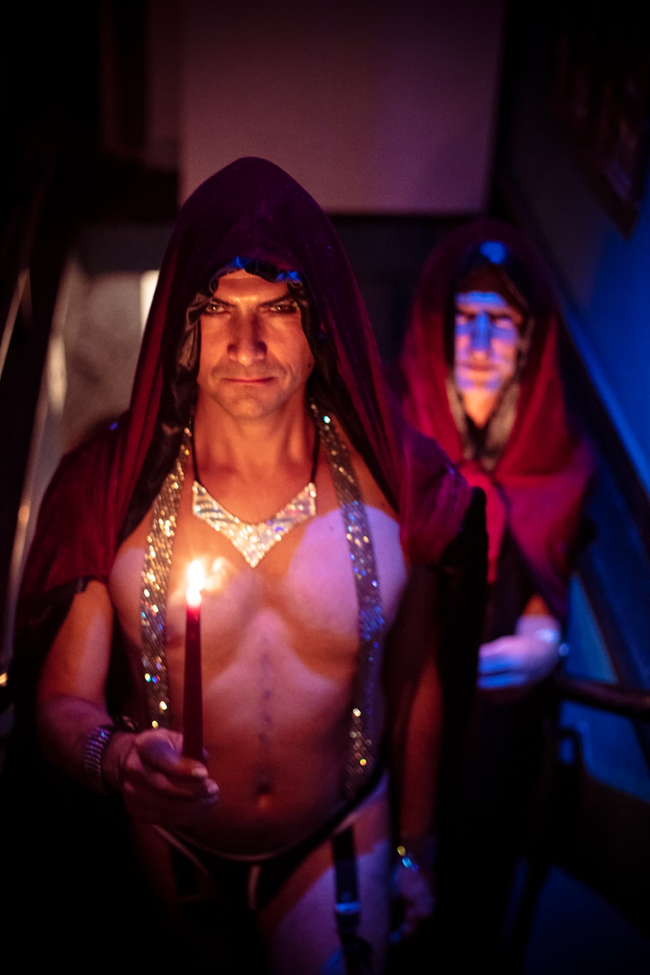 A camp member in a red hooded cape carrying a lit candle down a dark stairway, a second cloaked figure following behind