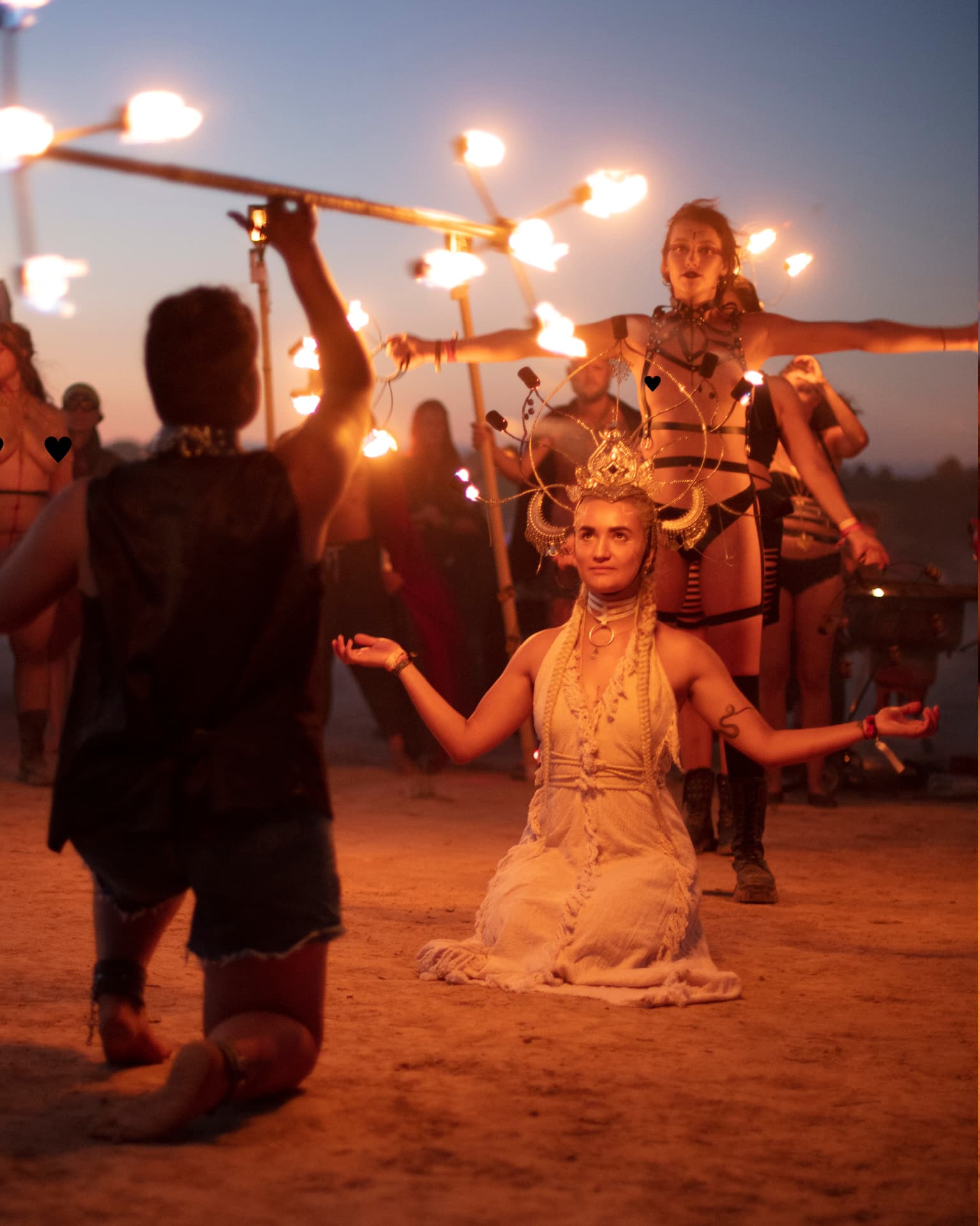 A kneeling performer in a golden crown surrounded by fire performers at dusk